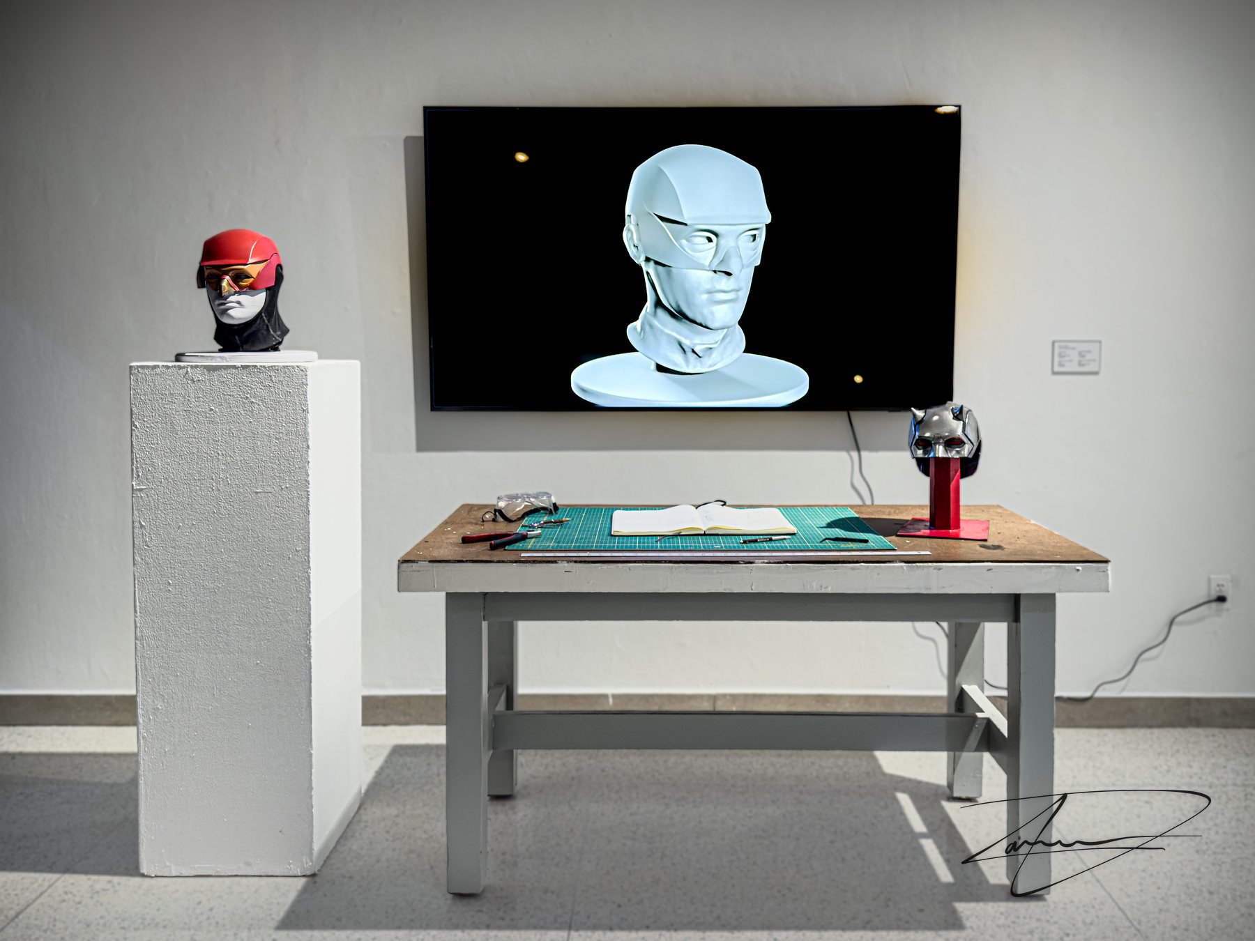 The installation as it was shown: the helmet on a pedestal, the workbench table carrying the sketchbook and the tools, and a monitor looping the progression from the head scan to the finished model.